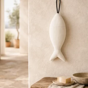 Olé & Co. Off-white linnen visje van gestoomd Europees linnen hangend aan kalkmuur in landelijke mediterrane ruimte, met olijfboom op de achtergrond en houten bank met linnen doek en zeep in zacht daglicht.